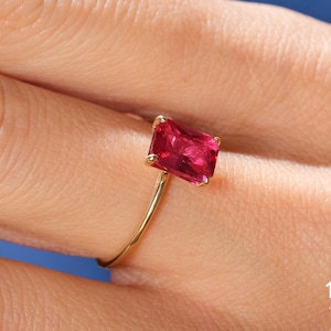 May include: A gold ring with a large, square-shaped, red gemstone. The ring is on a person's finger.