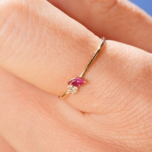 May include: A gold ring featuring a marquise-cut ruby and two small diamonds. The ring is set on a thin gold band. The ring is on a finger, with the brand name "1789 JEWELS" in the lower left corner.