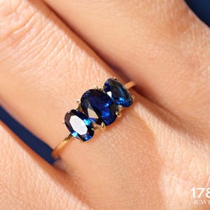 May include: A gold ring with three oval-shaped blue gemstones. The ring is on a finger.