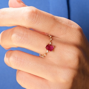 Puede incluir: Un anillo de oro con una piedra preciosa roja y pequeños diamantes. El anillo está en un dedo.