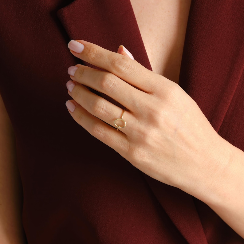 Puede incluir: Un delicado anillo de oro con un dise&ntilde;o ovalado abierto. El anillo se lleva en una mano con u&ntilde;as cuidadas, sobre un fondo burdeos. La joya es simple y elegante.