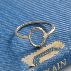 Puede incluir: Un delicado anillo de oro con un dise&ntilde;o de c&iacute;rculo abierto y una peque&ntilde;a gema transparente. El anillo se encuentra sobre un fondo azul, creando un contraste que resalta la elegante simplicidad del anillo. El dise&ntilde;o del anillo es moderno y minimalista.