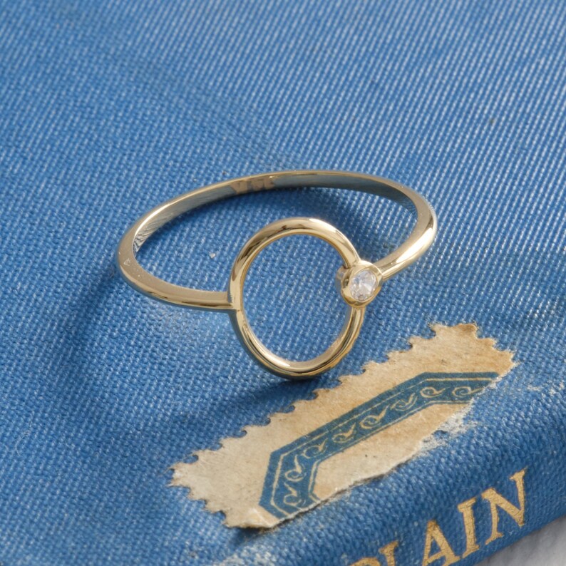 Puede incluir: Un delicado anillo de oro con un dise&ntilde;o de c&iacute;rculo abierto y una peque&ntilde;a gema transparente. El anillo se encuentra sobre un fondo azul, creando un contraste que resalta la elegante simplicidad del anillo. El dise&ntilde;o del anillo es moderno y minimalista.