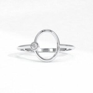 Puede incluir: Anillo de plata con dise&ntilde;o ovalado y una peque&ntilde;a gema redonda y transparente. El anillo tiene una est&eacute;tica minimalista y moderna, sobre un fondo blanco.