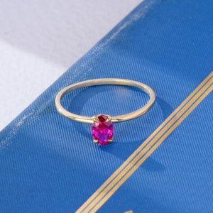 May include: A gold ring with a bright pink oval gemstone. The ring is on a blue surface.