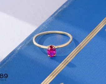 May include: A gold ring with a bright pink oval gemstone. The ring is on a blue surface.