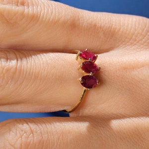 May include: A gold ring featuring three oval-cut, vibrant red gemstones set in a vertical line. The ring is displayed on a person's finger, with the brand name "1789 JEWELS" visible in the lower right corner.