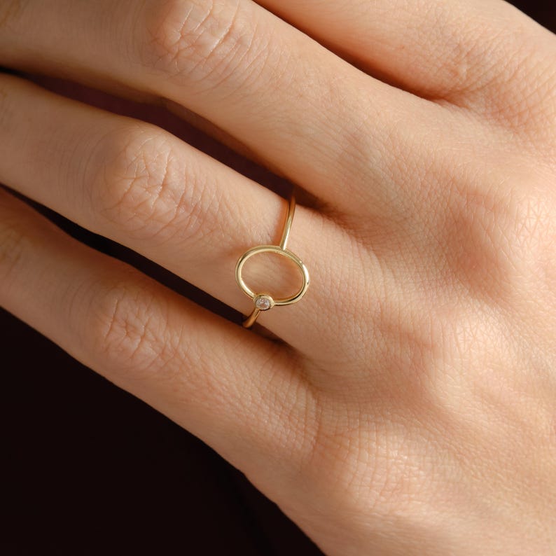 Puede incluir: Un delicado anillo de oro con una peque&ntilde;a gema transparente engastada en un dise&ntilde;o circular. El anillo tiene una banda delgada y elegante, con una est&eacute;tica minimalista. El anillo se muestra en el dedo de una persona.