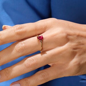May include: A gold ring with a red gemstone and two smaller diamonds. The ring is on a person's finger.