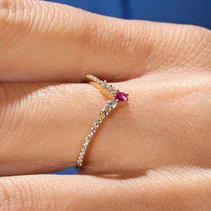May include: A V-shaped gold ring featuring a central round-cut pink gemstone flanked by small clear stones along the band. The band is set with multiple tiny white stones that extend along the chevron-style design. Text 1789 JEWELS is visible.