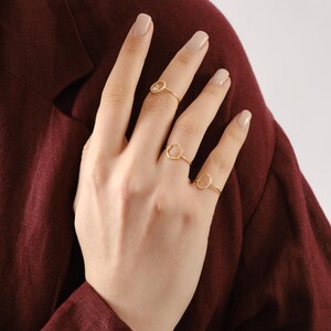 Puede incluir: Una mano con cuatro anillos dorados. Los anillos presentan una peque&ntilde;a piedra transparente, una forma hexagonal y una forma circular. El fondo es de color burdeos.