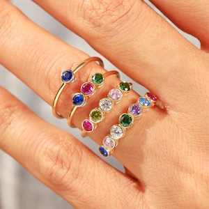 May include: A set of gold rings with colorful gemstones. The rings are stacked on a finger, with the gemstones arranged in a rainbow pattern.