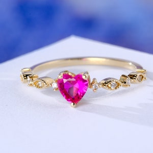 May include: A gold ring featuring a pink heart-shaped gemstone and small clear stones. The ring has a delicate, intricate design.
