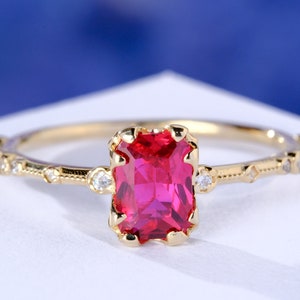 May include: A gold ring featuring a rectangular, vibrant red gemstone. The band is adorned with small, clear accent stones and decorative details. The ring is set against a white surface with a blue background. The text "1789 JEWELS" is visible.