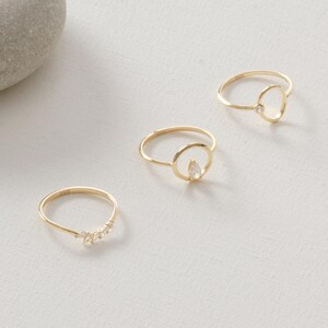 Puede incluir: Se muestran tres anillos dorados sobre una superficie blanca. Un anillo presenta una fila de peque&ntilde;as piedras transparentes, mientras que los otros dos tienen un dise&ntilde;o circular con una sola piedra transparente. Los anillos son delicados y minimalistas.
