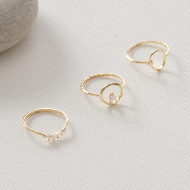 Puede incluir: Se muestran tres anillos dorados sobre una superficie blanca. Un anillo presenta una fila de peque&ntilde;as piedras transparentes, mientras que los otros dos tienen un dise&ntilde;o circular con una sola piedra transparente. Los anillos son delicados y minimalistas.