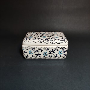 Pode incluir: Uma caixa de cerâmica branca com um design floral azul e preto. A caixa tem uma tampa com dobradiças e é decorada com um padrão repetitivo de flores e folhas estilizadas.