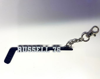May include: A hockey stick-shaped keychain in black and gray, with the name "RUSSELL" and the number "76" in gray lettering. The keychain includes a silver-colored clasp and chain.