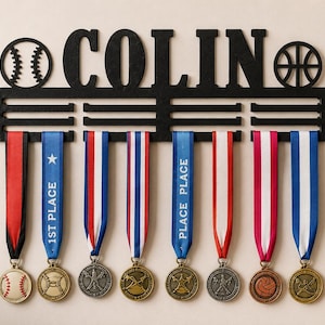 Könnte beinhalten: Ein schwarzer, wandmontierter Medaillenhalter mit dem Namen COLIN in großen Großbuchstaben, flankiert von einem Baseball- und Basketball-Symbol. Das Regal enthält drei horizontale Stangen zum Aufhängen von acht bunten Bändern mit Sportmedaillen.