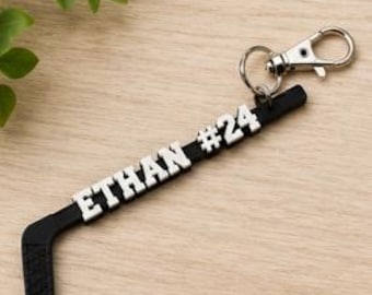May include: A black hockey stick keychain featuring the white raised text ETHAN #24 along the shaft. The keychain includes a silver-toned metal split ring and a lobster claw clasp attached to the top end of the hockey stick design.