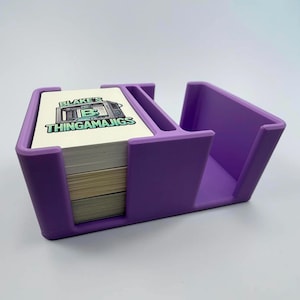 May include: A purple plastic card holder with two compartments. One compartment contains a stack of white rectangular cards featuring a graphic of a camera and the text Blake's Thingamajigs in green and black lettering. The second compartment is empty.