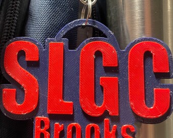 Könnte beinhalten: Ein blauer und roter 3D-gedruckter Schlüsselanhänger mit dem Text "SLGC Brooks" in roten Buchstaben.