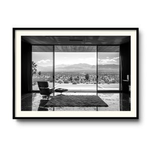 Pode incluir: Fotografia a preto e branco de um interior moderno com uma grande janela com vista para uma paisagem desértica. Uma poltrona e um pufe de couro preto estão colocados sobre um tapete em frente à janela. A imagem é emoldurada em uma borda preta.