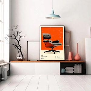 May include: A framed poster featuring a mid-century modern chair design. The poster has a black frame and a vibrant orange background. The chair is black and red with a black base. The poster is displayed on a white cabinet.
