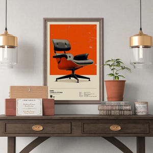 May include: A framed poster featuring an Eames Lounge Chair & Ottoman against an orange background. The chair is black and red, with a black base. The poster is displayed on a dark wooden table with a plant, books, and decorative items.