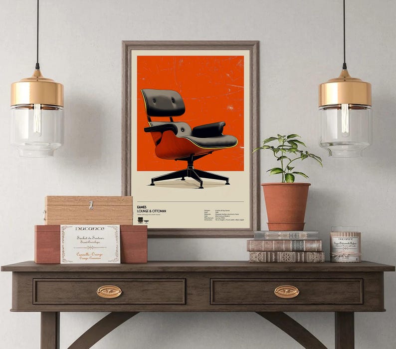 May include: A framed poster featuring an Eames Lounge Chair & Ottoman against an orange background. The chair is black and red, with a black base. The poster is displayed on a dark wooden table with a plant, books, and decorative items.