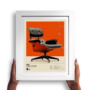 May include: Framed print of an Eames Lounge & Ottoman chair. The chair features a black leather seat and back with a wooden frame. The background is a vibrant orange, and the print is held in a white frame.
