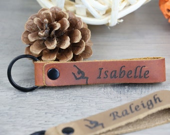 Op de afbeelding: Leren sleutelhangers met zwarte ringen. Eén sleutelhanger is bruin met de naam "Isabelle" en een turnster-silhouet. Een andere sleutelhanger is beige met de naam "Raleigh" en een turnster-silhouet.