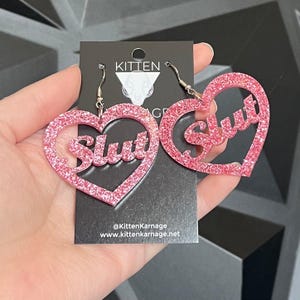 Op de afbeelding: Roze glitter hartvormige oorbellen met het woord "Slut" in een cursief lettertype. De oorbellen hangen aan zilverkleurige oorhaken. De oorbellen worden gepresenteerd op een zwarte kaart met het Kitten Karnage logo en website.