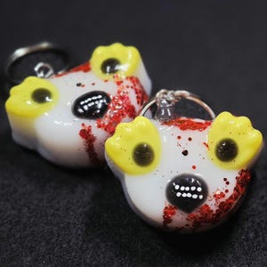May include: Two white bear-shaped keychains with yellow ears and black eyes and noses. The keychains have red glitter accents and silver key rings. The background is dark gray.
