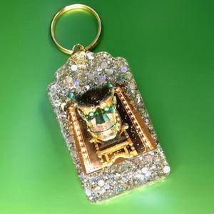 May include: A glittery keychain with a gold ring. The rectangular charm features a gold-colored frame with a detailed image of a figure on a chair, accented with green and gold. The charm is encased in clear resin with silver glitter.