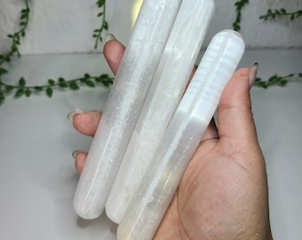 May include: Three white selenite wands, also known as selenite sticks, are held in a hand. The wands are smooth and have a striated texture.