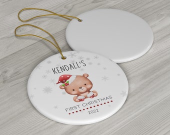 May include: White ceramic Christmas ornament with a cute cartoon bear wearing a Santa hat. The ornament says "Kendall's First Christmas 2022" and has a gold string for hanging.