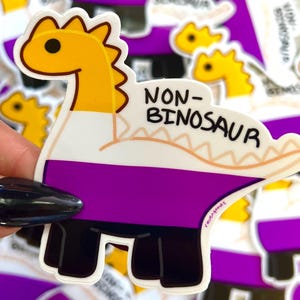 May include: A sticker featuring a dinosaur design with the text "NON-BINOSAUR". The dinosaur is colored with yellow, white, purple, and black, and has a cartoonish appearance. The sticker is held by a person.