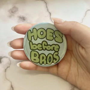 May include: A round, light blue button with the phrase "HOES before BROS" in green bubble letters. The button is held in a hand with long, manicured nails.