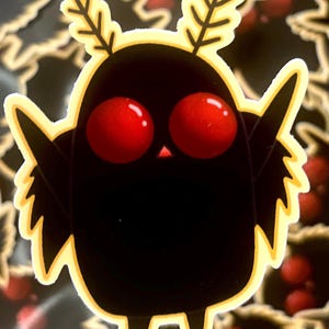 Könnte beinhalten: Ein schwarzer Cartoon-Kreatur-Aufkleber mit großen roten Augen, kleinen Flügeln und blattartigen Geweihen. Der Aufkleber hat einen gelben Umriss und ein festliches Design. Der Hintergrund ist dunkel mit goldenen und roten Akzenten.