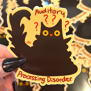 Può includere: Un adesivo raffigurante un gatto nero con occhi gialli e naso rosso. L'adesivo presenta le parole "Auditory" e "Processing Disorder" in rosso, con punti interrogativi sopra la testa del gatto. Il gatto è su sfondo giallo.