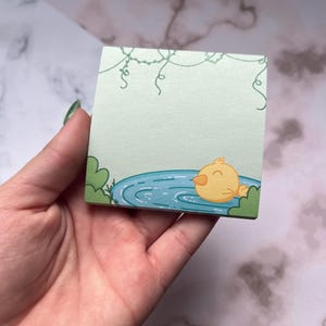 May include: A square sticky note pad with a light green background. The pad features a cartoon illustration of a yellow duck swimming in a blue pond. Green bushes and vines are also illustrated on the pad.