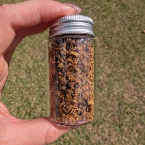 Könnte beinhalten: Ein kleines, klares Glas mit silbernem Deckel, gefüllt mit getrocknetem, zerkleinertem, braunem Pflanzenmaterial. Das Glas wird in der Hand einer Person vor dem Hintergrund von grünem Gras gehalten.
