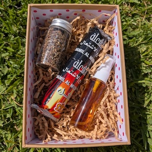 Könnte beinhalten: Eine Geschenkbox mit gepunktetem Innenraum enthält drei Artikel: ein Glas mit silbernem Deckel, gefüllt mit dunklen Granulaten, eine schwarz-rote Verpackung mit arabischem Text und eine transparente Sprühflasche mit bernsteinfarbener Flüssigkeit.