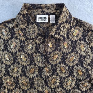 Può includere: Camicia nera e oro con motivo floreale in pizzo e dettagli di paillettes. La camicia ha un colletto e l'etichetta "Chico's Design".