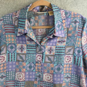 May include: A button-down shirt with a geometric pattern in shades of purple, teal, and peach. The shirt is hanging on a wooden hanger. The pattern features squares and rectangles with various shapes.