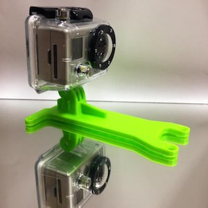 Puede incluir: Una cámara GoPro plateada con una lente negra y una carcasa protectora transparente está montada en un soporte de plástico verde brillante.