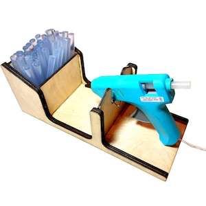 Puede incluir: Un soporte de madera con un compartimento para barras de pegamento y un soporte para una pistola de pegamento azul. La pistola de pegamento está enchufada y tiene un cable blanco.