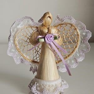 May include: A decorative angel figurine crafted from straw, featuring heart-shaped wings with a delicate lace trim. The angel has a lavender ribbon and rose detail, with a matching lace trim at the base. The overall design is light and airy.