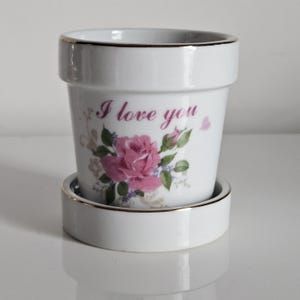 Puede incluir: Maceta de cerámica blanca con platillo a juego. La maceta presenta un diseño de rosa rosa y las palabras "I love you" en escritura cursiva. El borde del tiesto y del platillo están adornados con ribetes dorados. Un artículo decorativo para el hogar o el jardín.